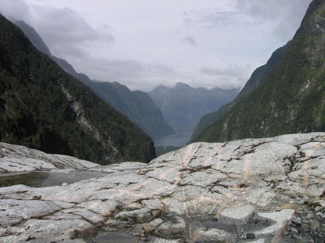 Figure 4. View toward Milford Village from Pembroke Valley, Fiordland. The Pembroke Granulite in foreground displays a rectilinear array of white to gray leucosomes and pink garnet reaction zones indicative of partial melting and wall rock reactions in the lower crust of the Cretaceous Gondwana Magmatic Arc. Photo credit: Harold Stowell.